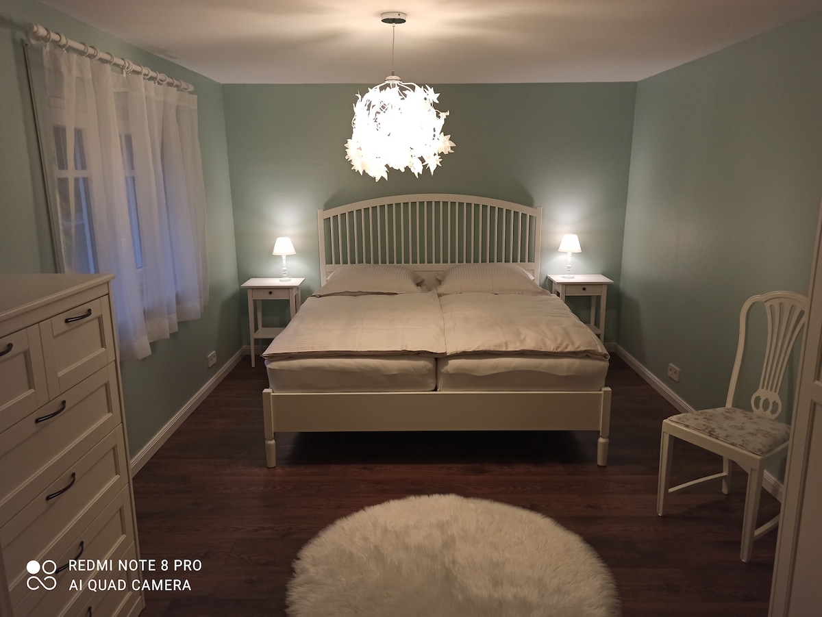 A softly lit bedroom features a spacious bed with light bedding and matching nightstands. A delicate chandelier provides illumination, while a light-colored chair adds a touch of elegance. A round rug lies beneath the bed, and sheer curtains frame the window, allowing natural light to shine through.