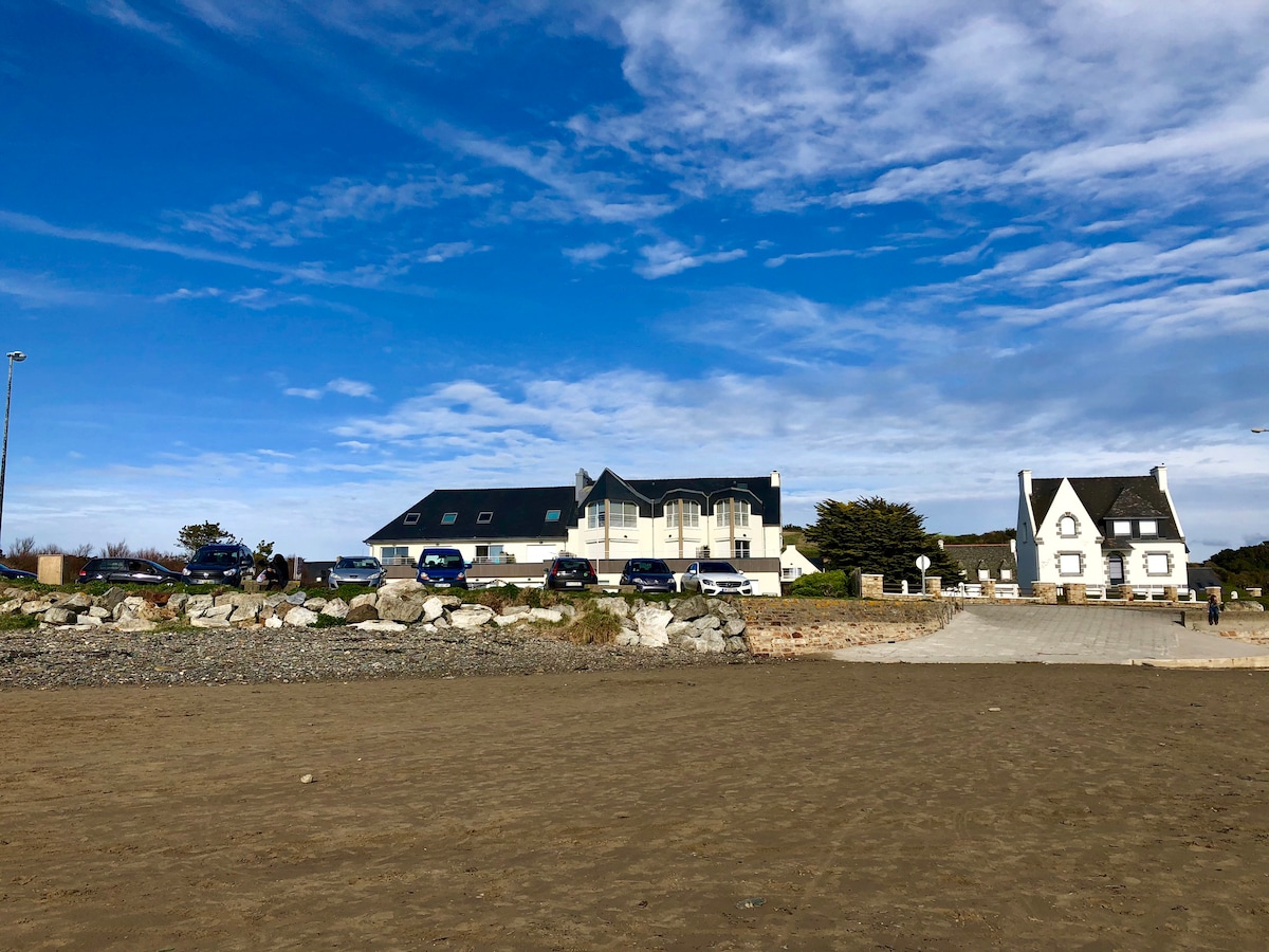A sandy beach leads up to a residence featuring two distinct buildings. The main structure has multiple windows, surrounded by a maintained area with several parked vehicles. The sky above is partly cloudy, contributing to the serene coastal atmosphere.