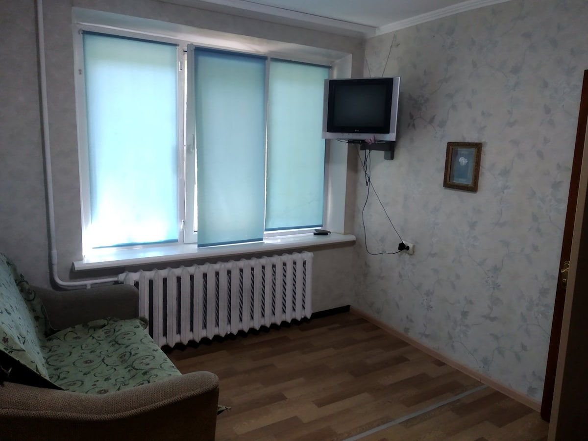 A room featuring a sofa with patterned upholstery is positioned near large windows covered with light blue blinds. A small wall-mounted television is installed opposite the windows, above a heating radiator. The walls are adorned with a subtle floral pattern.