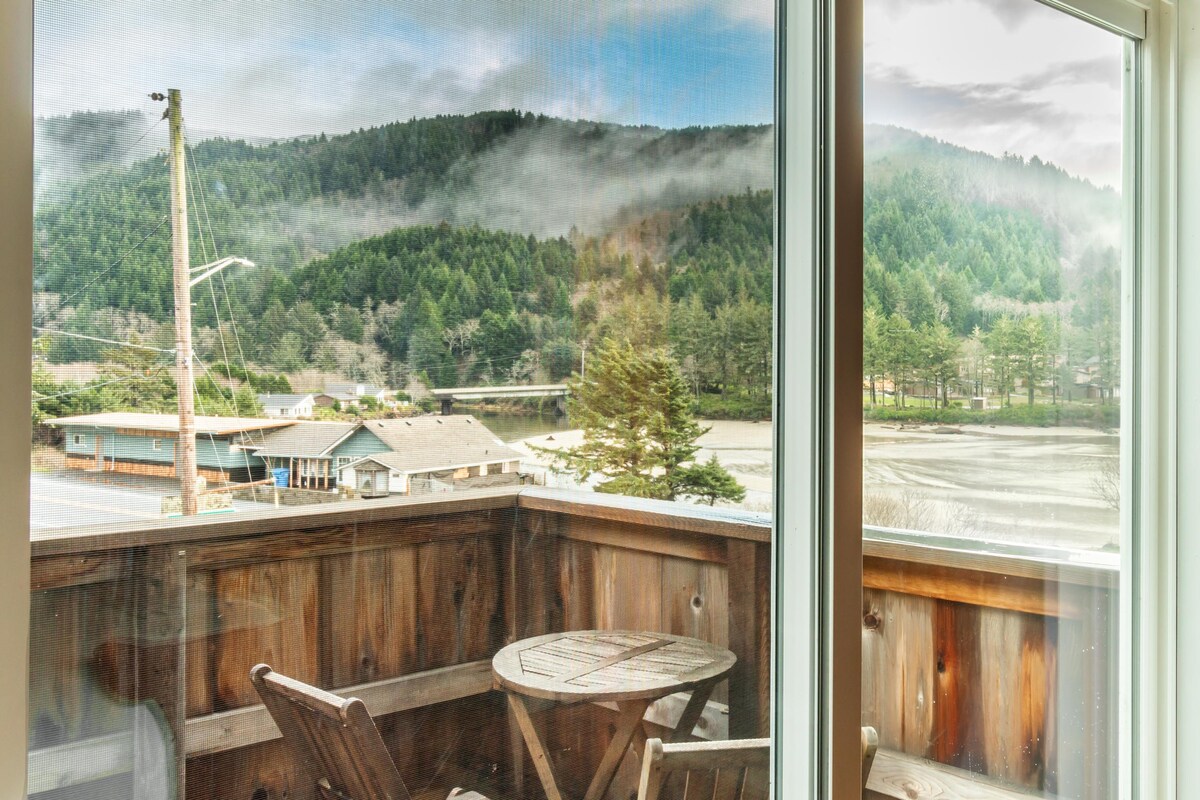 A view from the studio's deck showcases rolling green hills shrouded in mist. The landscape features a serene river winding through the area and modest structures nestled below against the backdrop of trees.