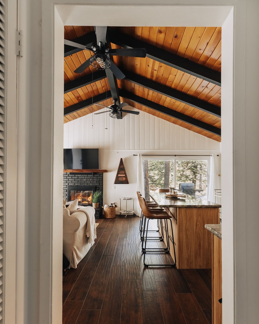 A view through a doorway reveals the main living area of the cabin, featuring high wood-vaulted ceilings and an inviting layout. A cozy couch is positioned in front of a gas fireplace, with a kitchen island and bar seating visible in the background, illuminated by natural light.