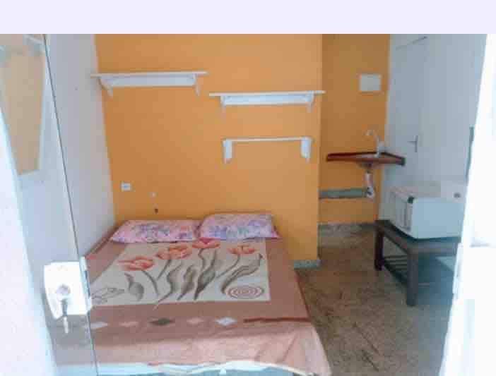 A simple room is presented with an orange accent wall. A double bed adorned with light floral bedding occupies the space, complemented by open shelves for storage. A small table is positioned beside the bed, and a kitchenette area is visible in the corner.