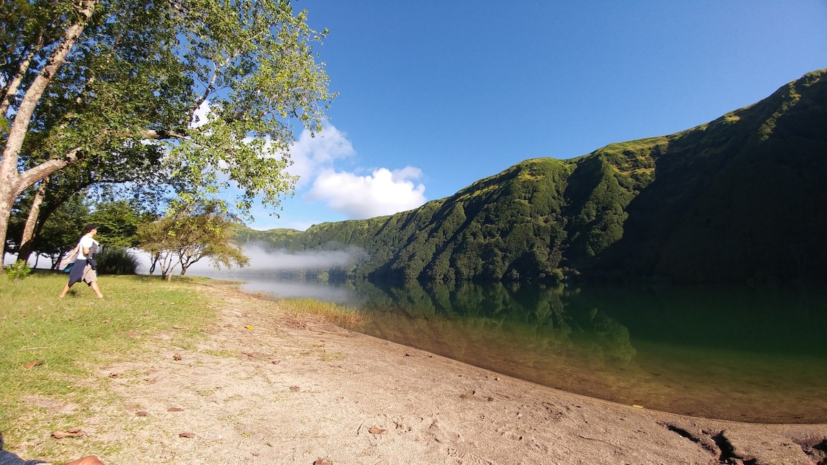 A serene lakeside view is depicted, featuring lush green hills reflected in still water. Nearby, patches of sandy shore are framed by trees, and soft, white clouds hover over the landscape. The scene captures a peaceful natural setting, inviting tranquility and relaxation.