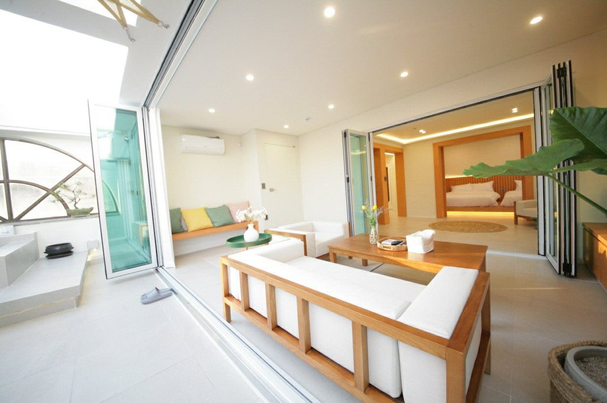 A bright living space is featured, with large glass doors opening to the outdoors. A comfortable seating area with light-colored cushions is complemented by a wooden coffee table. A plant adds a touch of greenery, while soft lighting creates a welcoming ambiance.