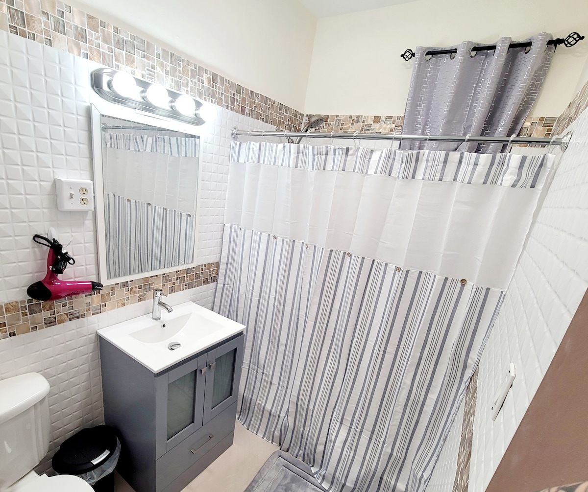 A well-appointed bathroom features a shower with a striped curtain and a modern sink vanity with storage. A clear mirror is positioned above the vanity, and a hairdryer is mounted on the wall. Natural light illuminates the space, complemented by decorative tiles and a neutral color palette.