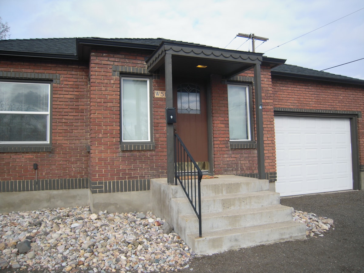 The entrance features a covered porch with a decorative overhang. A set of stairs leads up to a wooden front door, framed by two large windows. A one-car garage is visible to the right, and gravel landscaping surrounds the base of the home.
