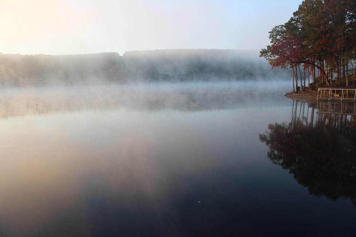 A serene lake view is captured, showcasing calm waters reflecting the surrounding trees and distant hills. Morning mist hovers over the lake's surface, creating a tranquil atmosphere. The shoreline features a small wooden dock surrounded by trees with hints of autumn color.