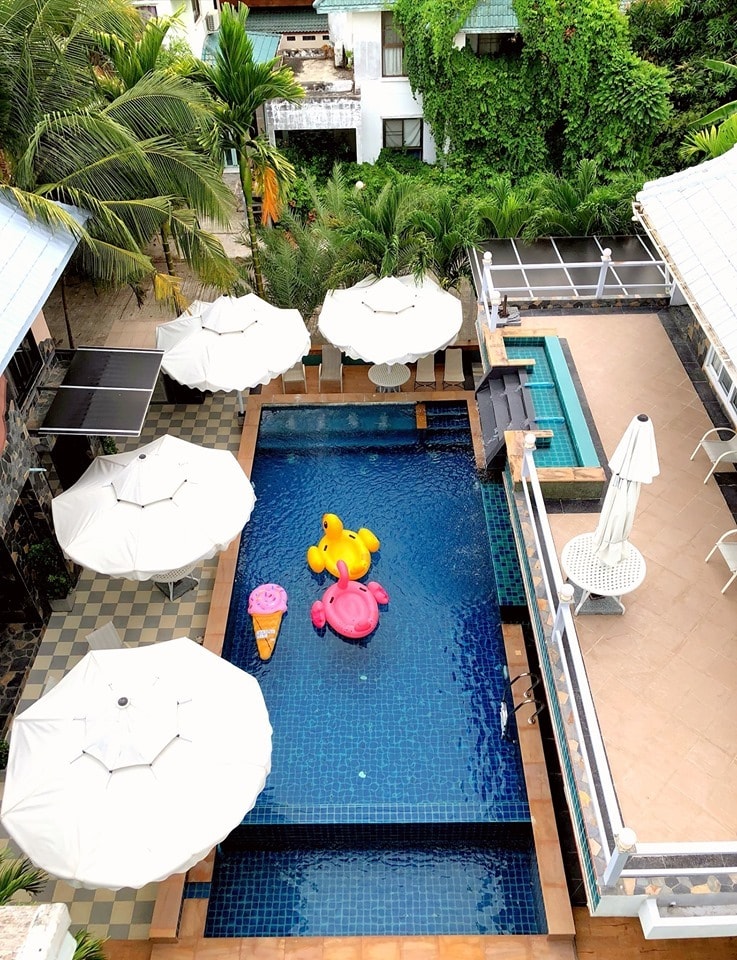 The aerial view captures a large private pool surrounded by umbrellas and tropical greenery. Colorful pool floats are visible in the water, complementing the inviting blue tiles. Adjacent to the pool, a jacuzzi area features an elegant seating arrangement.
