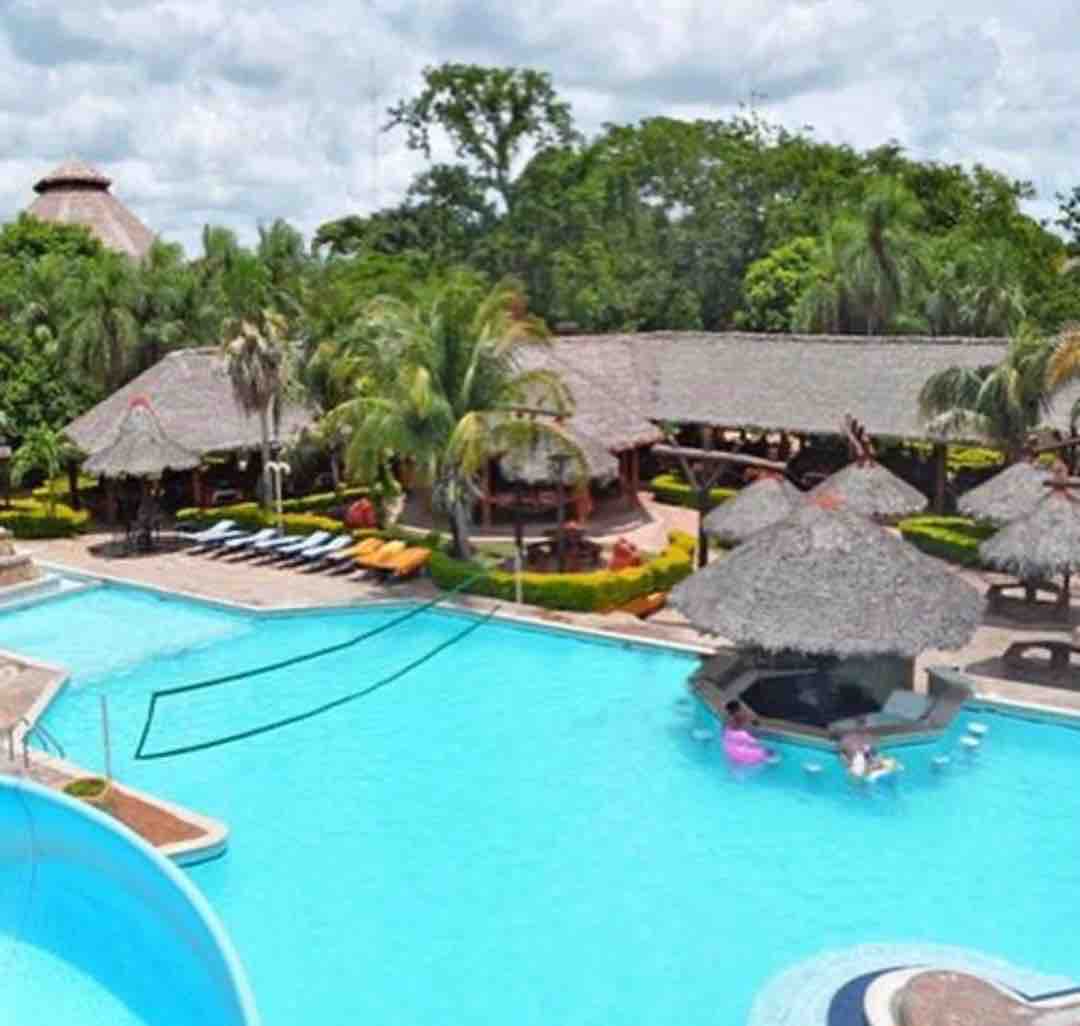 A large swimming pool is surrounded by lush greenery and tropical palms. Thatched-roof cabanas and seating areas are visible, offering shaded space for relaxation. The clear blue water glimmers under the daylight, creating a serene atmosphere for leisure activities.