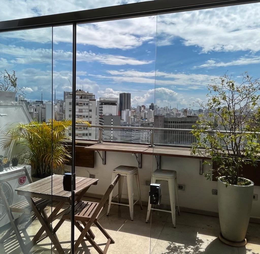 A spacious balcony offers a striking view of São Paulo's skyline, framed by glass railings. A wooden table and stools provide seating, while potted plants add a touch of greenery. The sky is bright, with clouds scattered across a blue backdrop.
