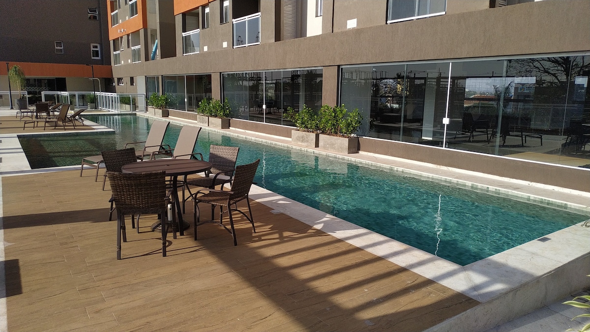 A tranquil outdoor pool area features a clear water pool surrounded by lounge chairs and a dining table with four woven chairs. Potted plants are placed along the poolside, enhancing the inviting atmosphere. Large glass windows of the building reflect the surroundings.