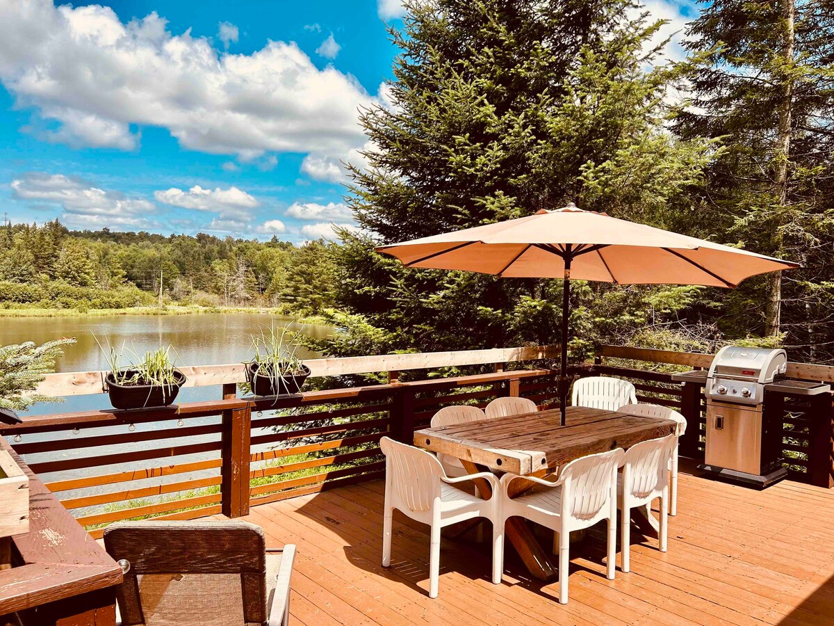 An outdoor deck features a large wooden dining table surrounded by white chairs, with a large umbrella providing shade. Potted plants add greenery, and a barbecue grill is visible nearby, all set against a serene lake view and lush trees.