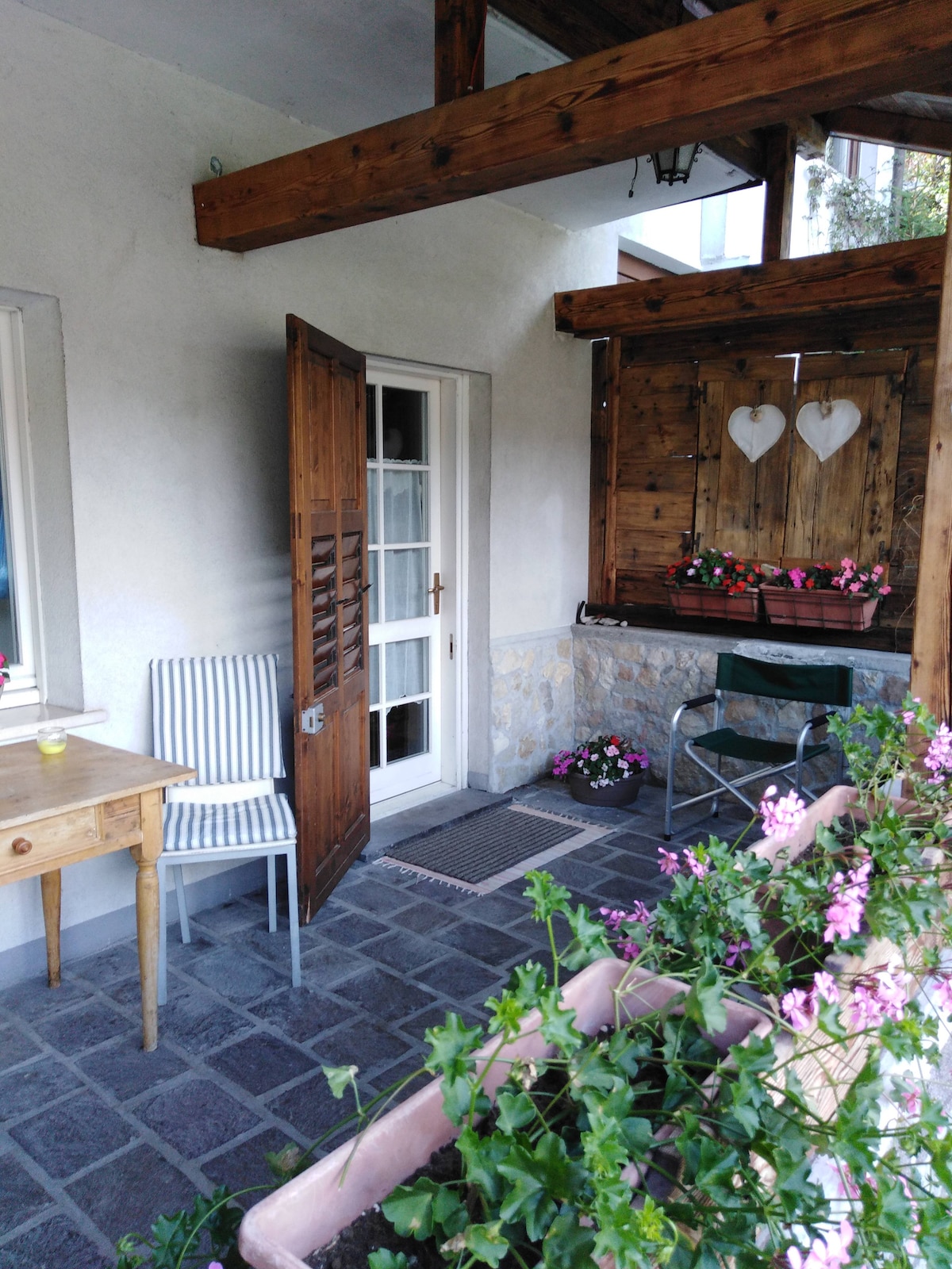 An inviting outdoor porch area is featured, complete with a small wooden table and a striped chair. A door leads to the interior, while vibrant flowers in containers add color to the space, enhancing the tranquil setting.