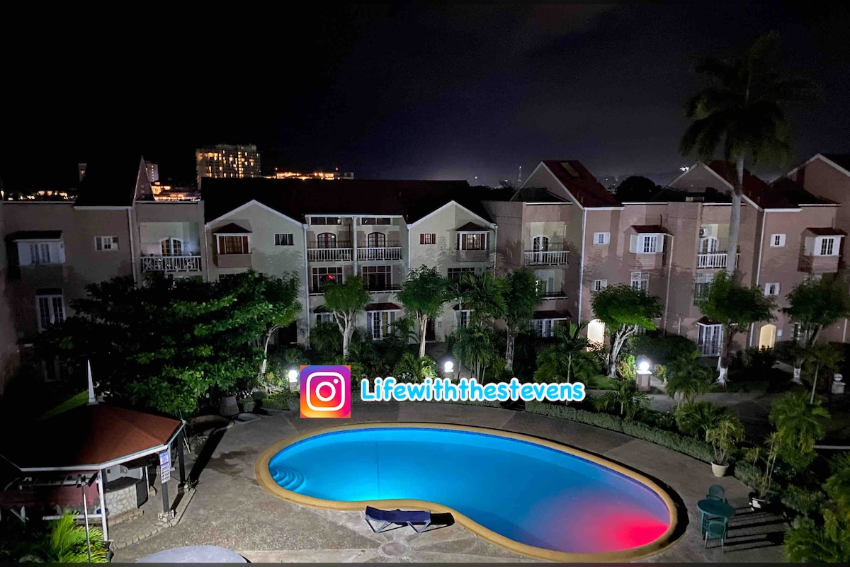 An oval-shaped pool, illuminated in blue and red, is surrounded by a spacious courtyard featuring tropical landscaping. Nearby, several buildings with balconies are visible, reflecting a calm nighttime atmosphere, while palm trees add a touch of greenery.