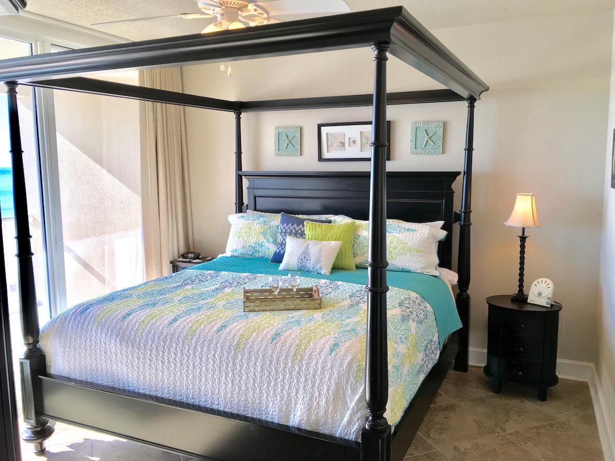 A four-poster bed with a colorful quilt draws attention, surrounded by soft neutral walls and natural light from expansive glass doors. A bedside table with a lamp is positioned beside the bed, adding functionality to the room's serene layout.