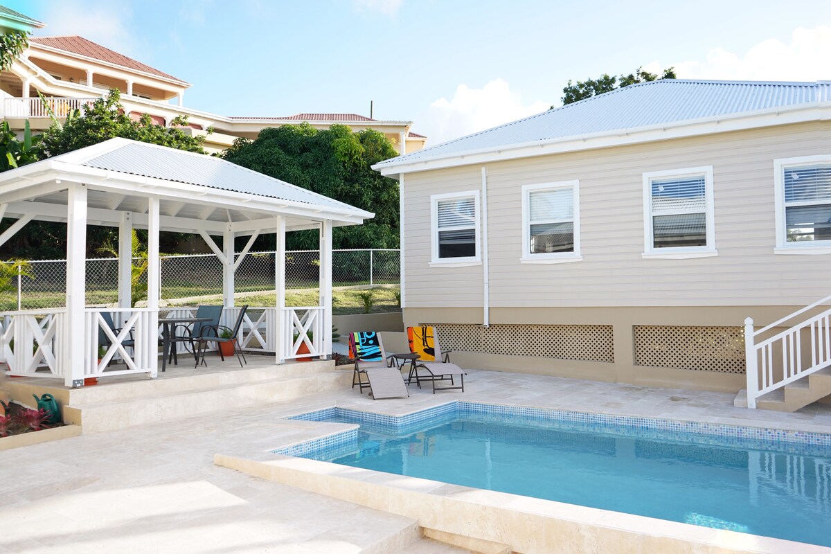A well-maintained patio area features a swimming pool surrounded by sun loungers. A shaded gazebo with a seating arrangement provides a relaxing spot. The exterior of the cottage is light-colored, and windows allow natural light into the interior.