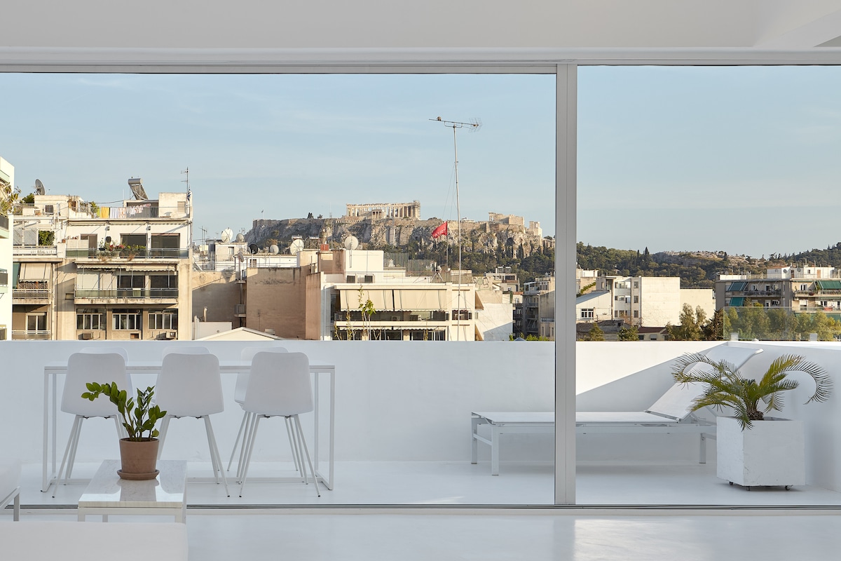 A modern terrace overlooks the Acropolis, framed by large glass doors. Two white chairs and a small potted plant are positioned around a minimalist table, with a sunbed adjacent, creating a refreshing outdoor space bathed in natural light.