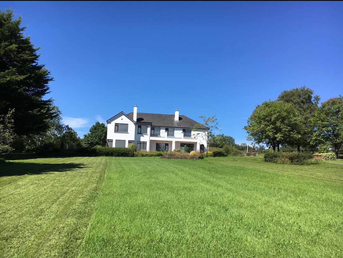 A spacious two-story house is shown against a clear blue sky, framed by lush green grass and trees. The property features a well-maintained lawn and a blend of landscaped shrubbery surrounding the home.