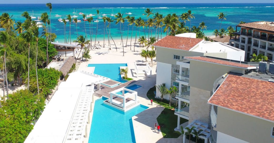 An aerial view showcases a large swimming pool surrounded by palm trees, leading to a sandy beach and the ocean beyond. The vibrant blue waters reflect sunlight, and nearby boats are seen on the horizon. Elegant buildings dot the landscape, enhancing the beachfront setting.