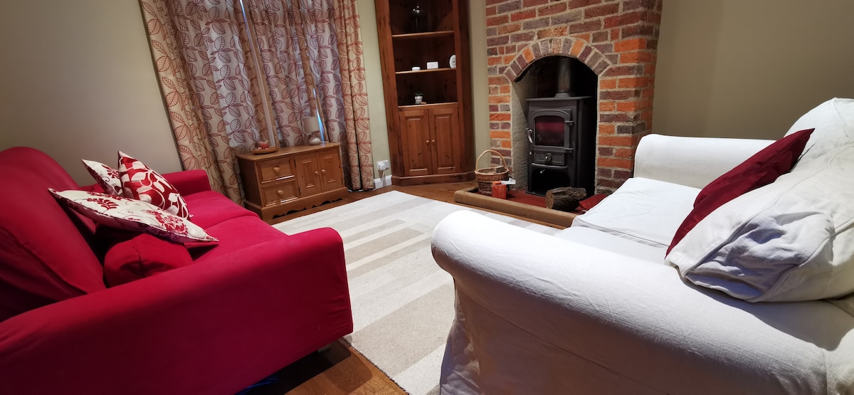 A cozy living area features a white sofa and a red sofa adorned with patterned cushions. A warm brick fireplace adds character to the space, while a light area rug and curtains create a gentle ambiance.