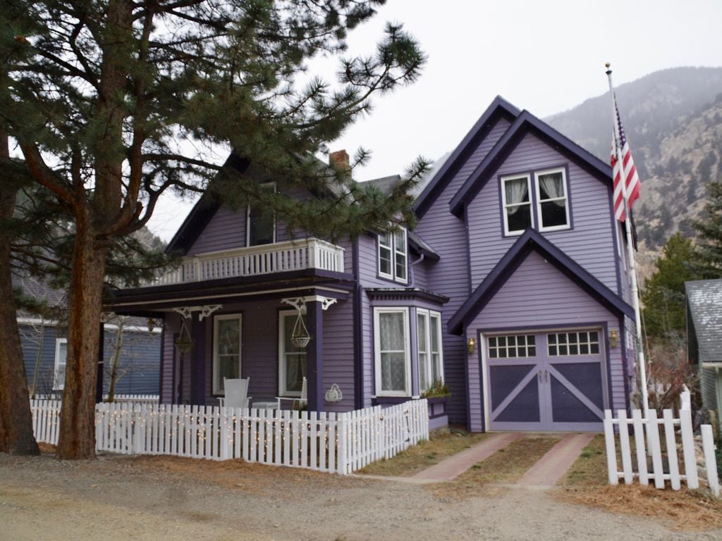A charming Victorian house is showcased, featuring a distinct purple exterior. A covered porch is visible, along with decorative railings and a small front garden. An American flag is displayed, and a driveway leads to a garage. Tall pine trees frame the house against a mountainous backdrop.