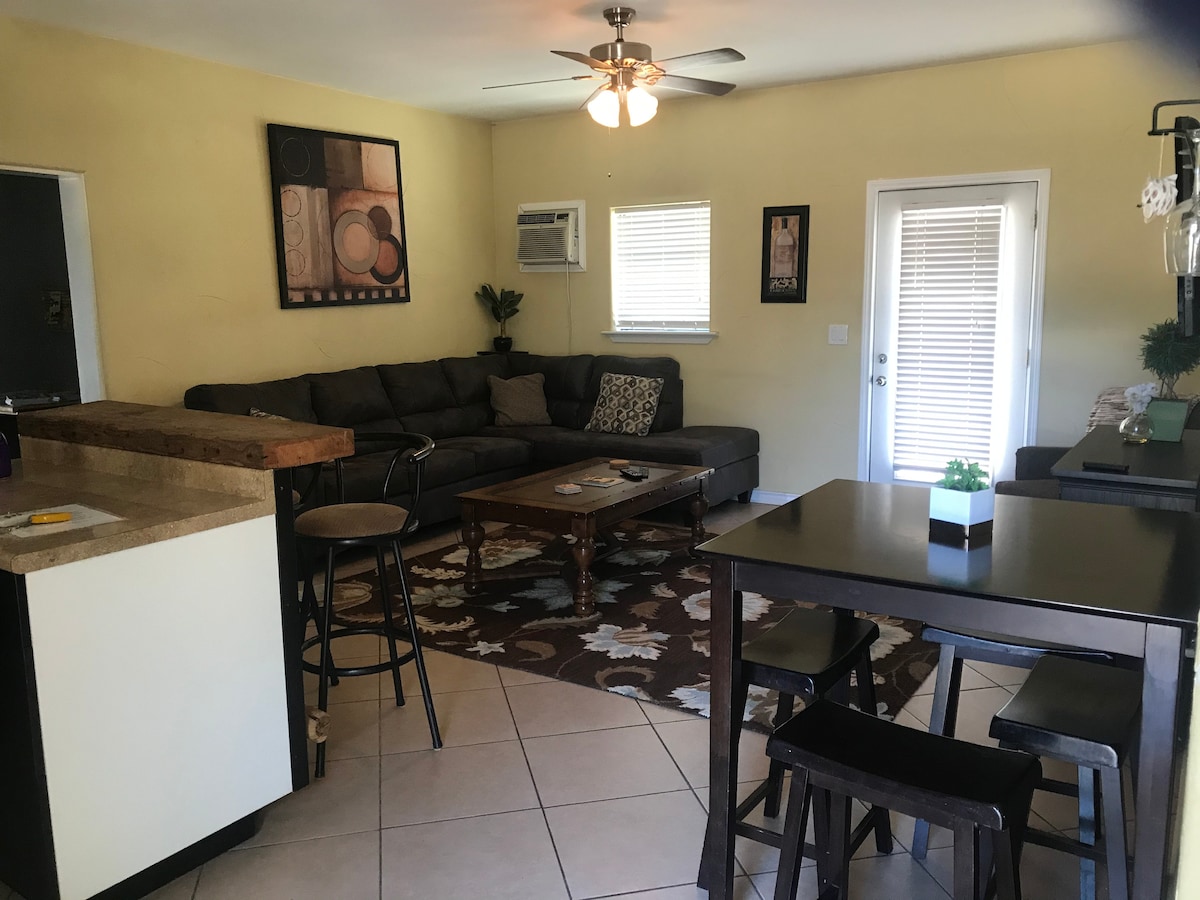 A bright living area features a comfortable sectional sofa centered on a patterned area rug. A small coffee table is positioned in front, and a dining table with seating for four is visible. Natural light enters through a nearby door with blinds.