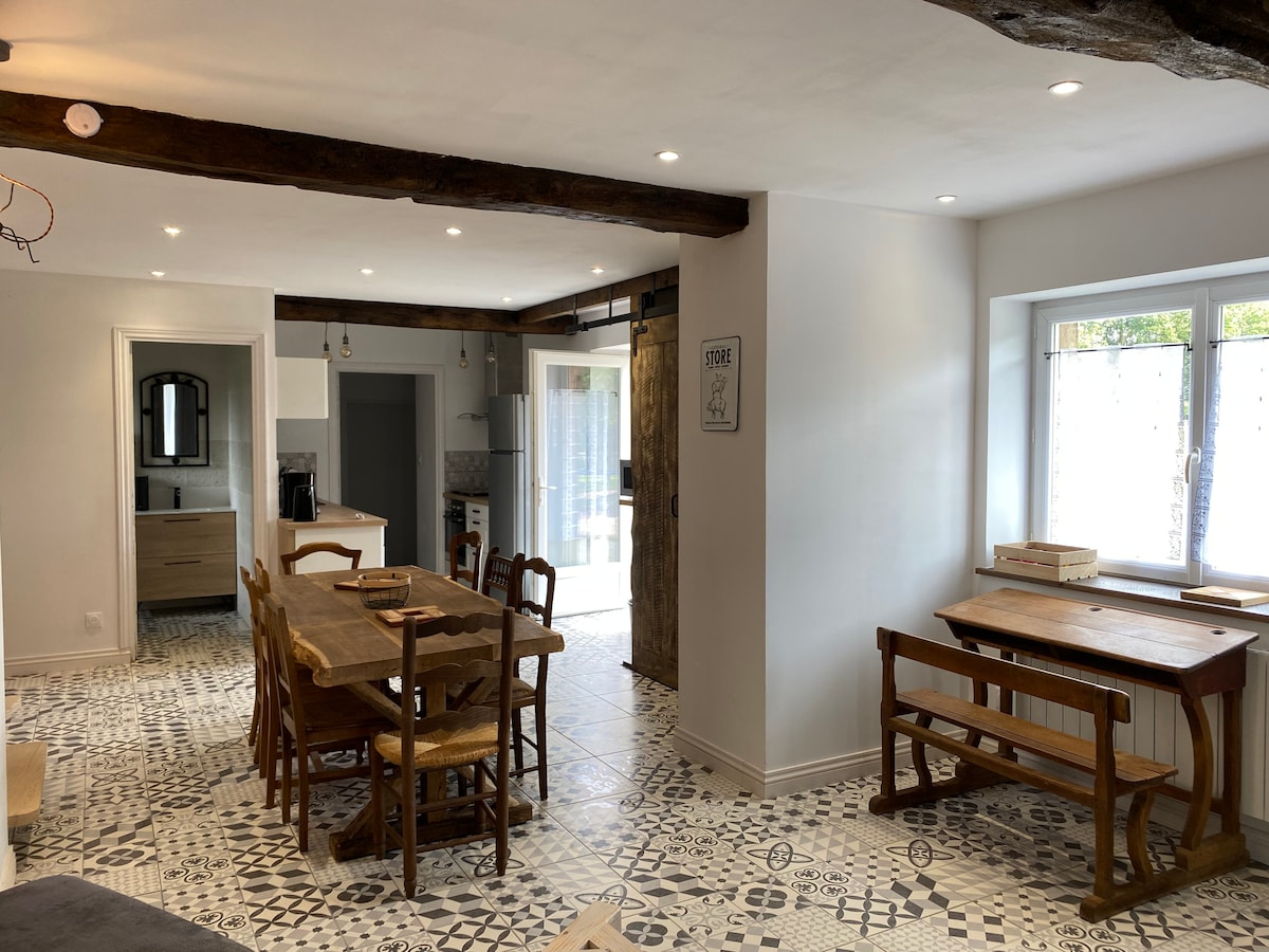 The open-concept living area features rustic wooden beams and patterned tiled flooring. A large dining table with chairs is positioned adjacent to a fully equipped kitchen. Natural light pours in through windows, creating a bright and inviting atmosphere in the communal space.