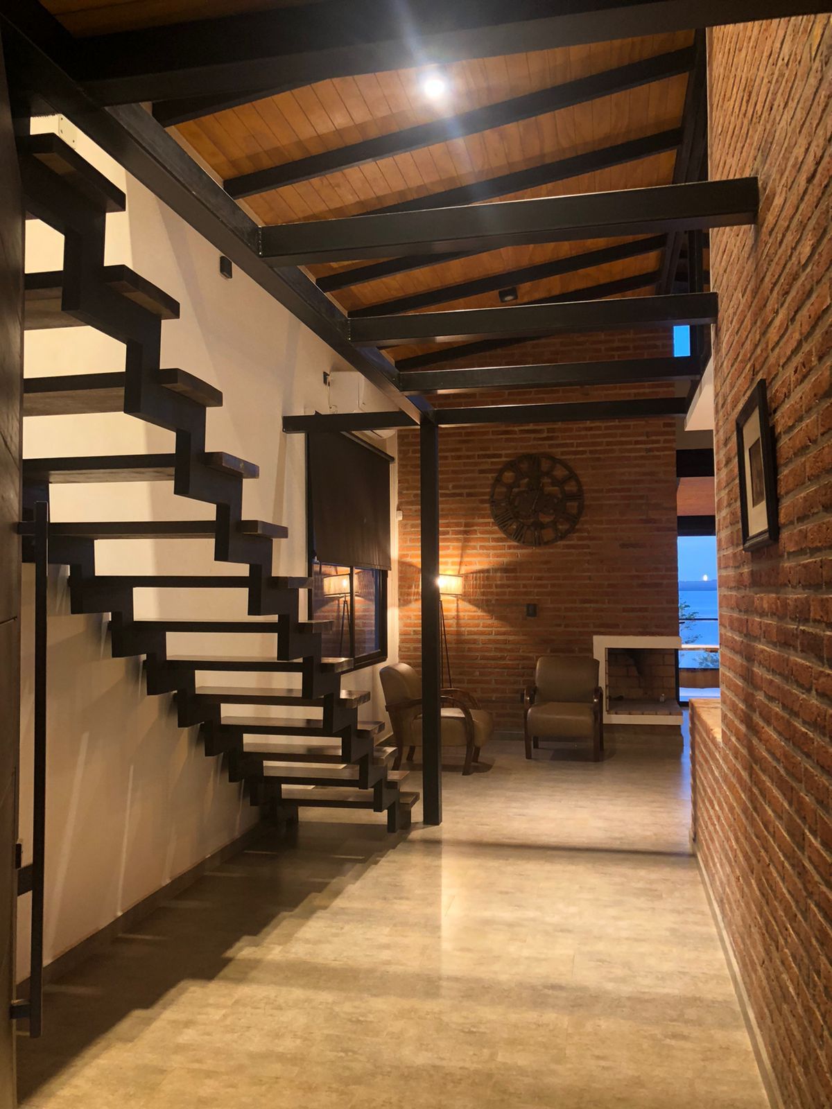 An open space features a modern staircase with a sleek design leading to the upper level. Exposed brick walls and wooden beams add a rustic touch, while a pair of armchairs is positioned nearby, creating a cozy area for relaxation. Large windows provide a glimpse of the outdoor scenery.
