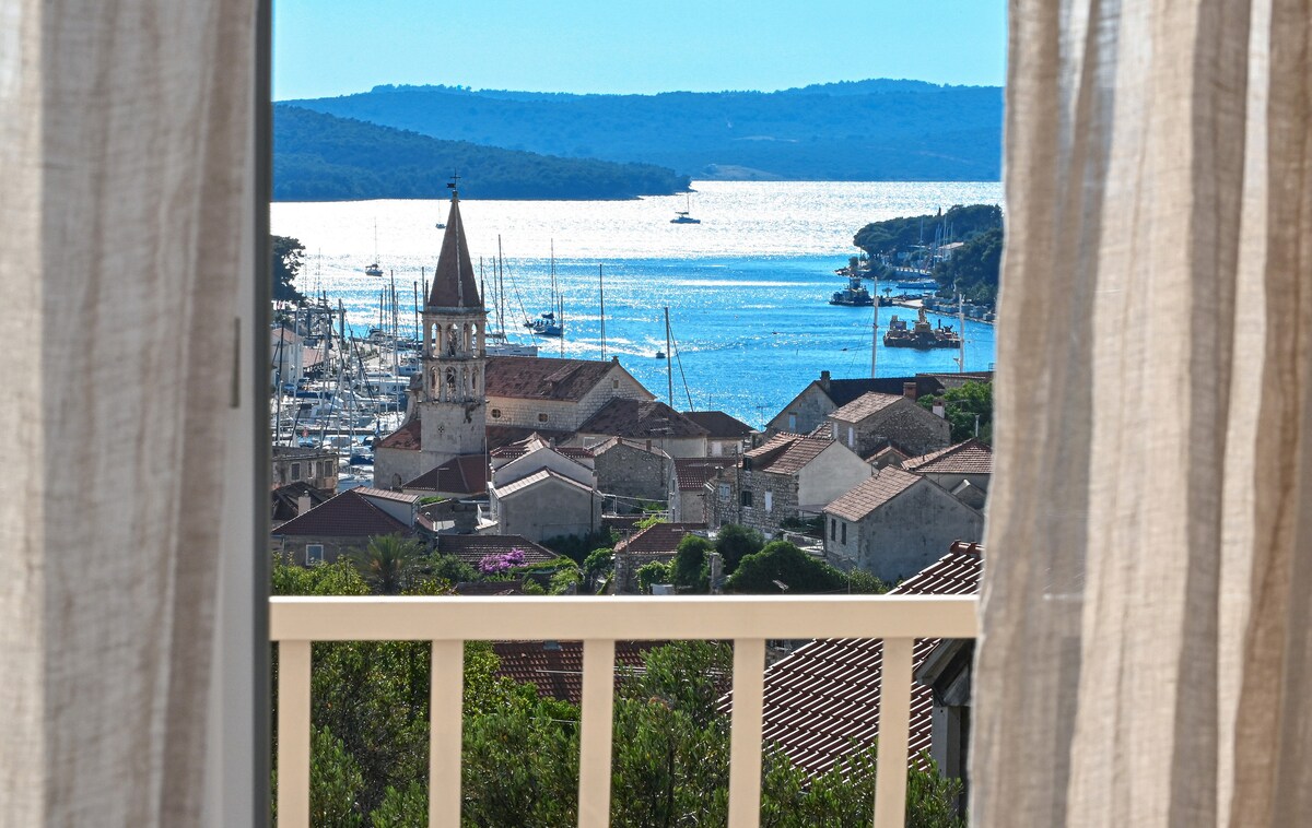 A scenic view is framed by soft, sheer curtains, showcasing the tranquil harbor with sailboats and the charming town below. The distant outline of hills is visible in the background, enhancing the peaceful coastal atmosphere.