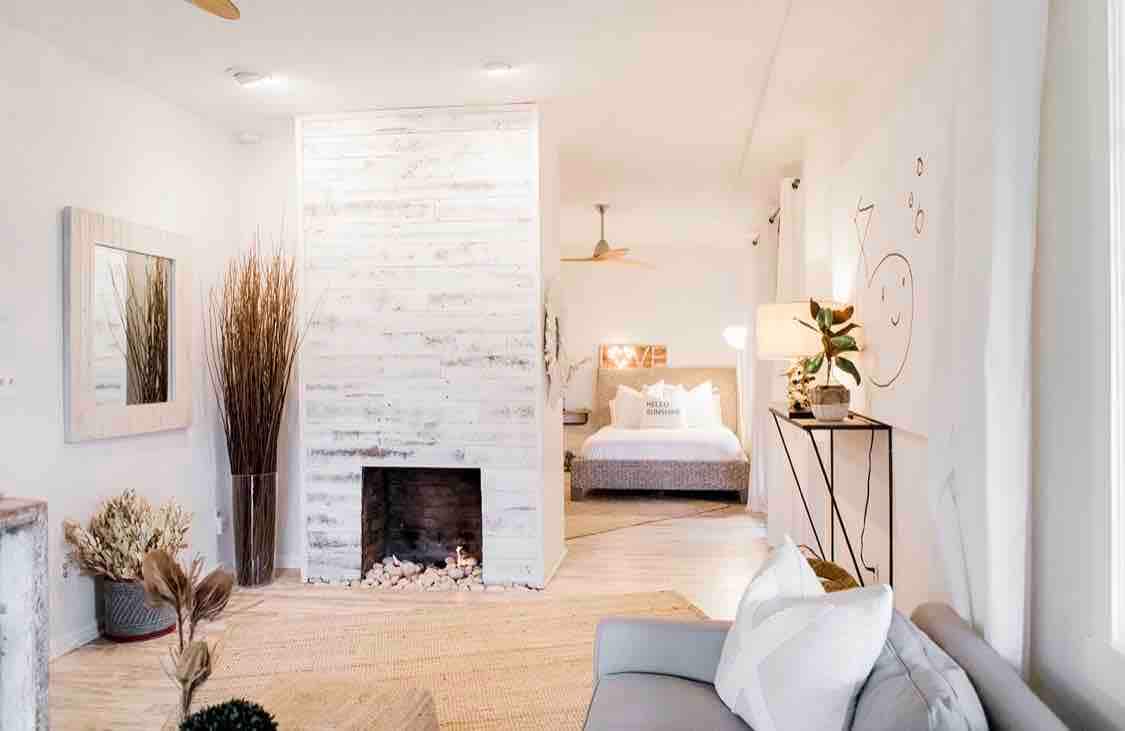 A comfortable living space is shown with a neutral color palette, featuring a modern sofa and decorative plants. A distinctive whitewashed fireplace serves as a focal point, while a bedroom area is visible in the background, enhanced by natural light from nearby windows.