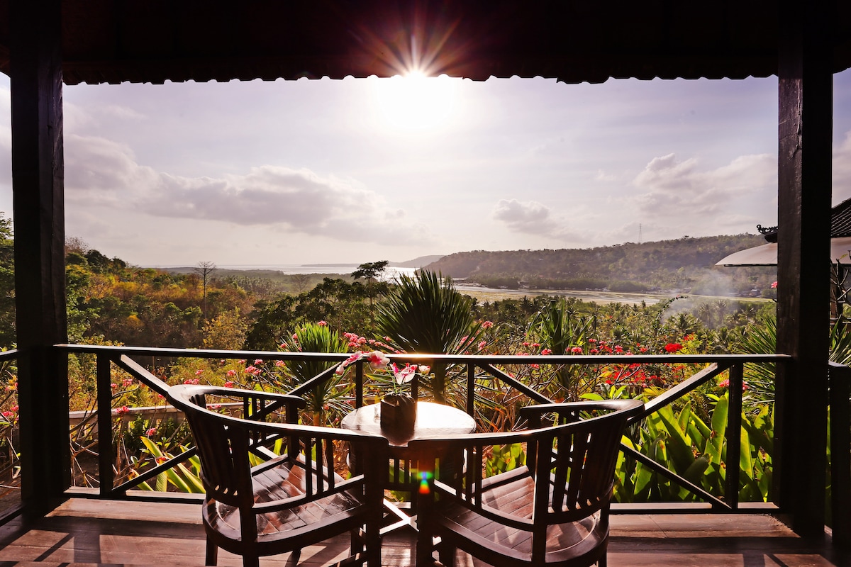 A wooden table with two chairs is positioned on a private terrace, offering a panoramic view of lush greenery and hills. The sun rises over the landscape, casting a warm glow, while colorful flowers frame the scene. Tranquility and natural beauty are emphasized.