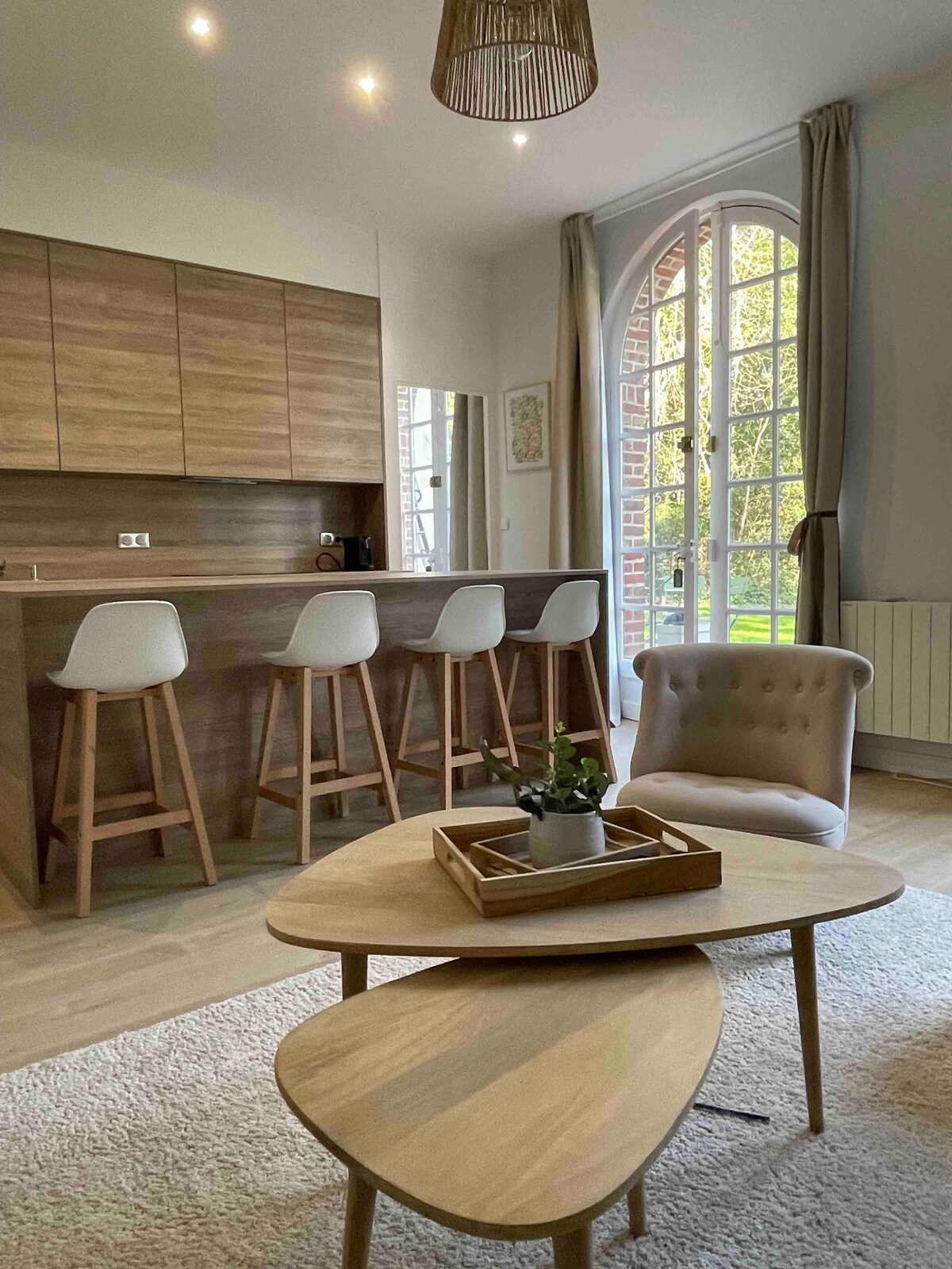 A welcoming kitchen area features a wooden bar with four stools. Natural light floods through large windows, illuminating the space. A stylish, light-colored armchair sits near a pair of modern wooden coffee tables, complemented by a small plant centerpiece.