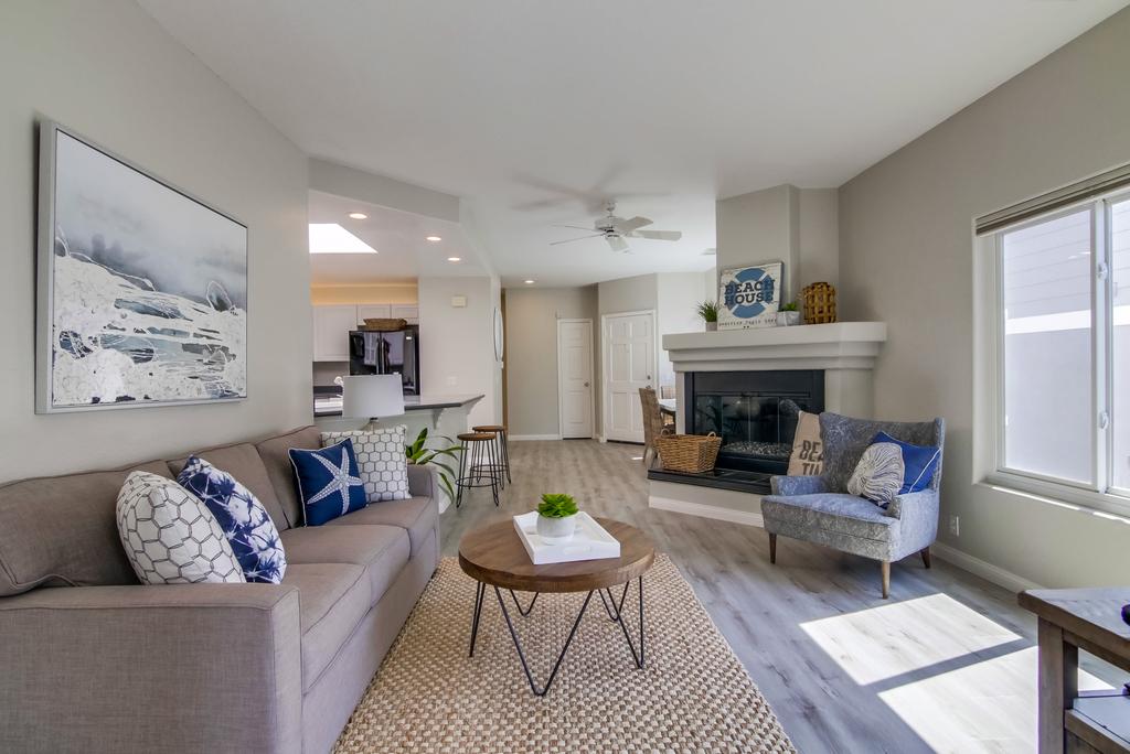 A spacious living room presents a comfortable sofa adorned with decorative pillows. A coffee table with a plant sits centrally. A gas fireplace is positioned against a light-colored wall, while a ceiling fan adds an airy feel. Natural light enters through a nearby window.