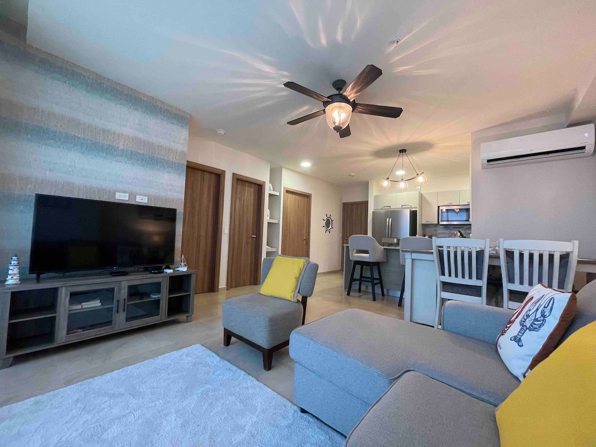A modern living room features a comfortable gray sofa with a colorful throw pillow, positioned near a light-colored area rug. A flat-screen television is mounted on the wall, while a dining area with light wooden chairs and a kitchen can be seen in the background.