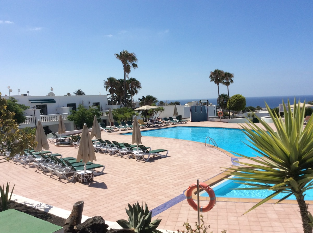 A large swimming pool is framed by sun loungers and umbrellas on a tiled deck. Palm trees are visible in the background, and a view of the sea can be seen beyond the pool area, under a clear blue sky.
