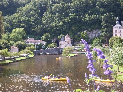 A serene river scene is depicted, with several kayaks gently gliding across the water. Lush greenery frames the landscape, with trees and shrubs lining the banks. A charming stone structure is visible in the background, alongside riverfront pathways and reflections on the water.
