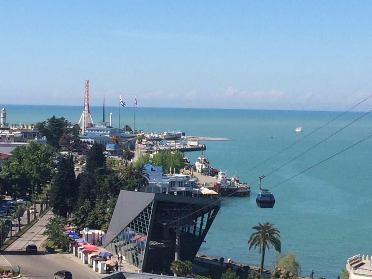 This image captures a panoramic view of the seaside, featuring a vibrant waterfront area. A cable car is visible moving toward hills in the background. The coastline is dotted with boats, palm trees, and various structures, including recreational areas and amusement attractions.