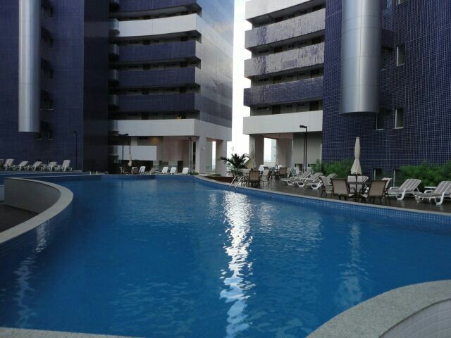A large outdoor swimming pool is framed by modern buildings, featuring lounge chairs positioned along the edge. The water reflects the surrounding architecture, and palm trees are visible in the background, enhancing the open, inviting atmosphere.