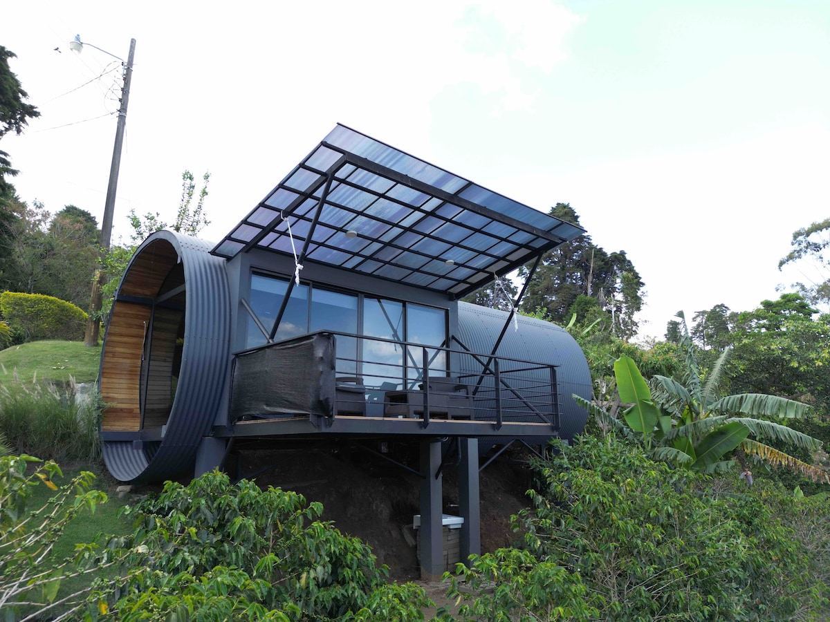 A unique rural retreat is captured, featuring a modern cylindrical structure with large glass windows and a distinctive pointed roof. A balcony extends outward, offering a view of the surrounding greenery and coffee plantations. Solar panels are positioned atop the structure for sustainability.