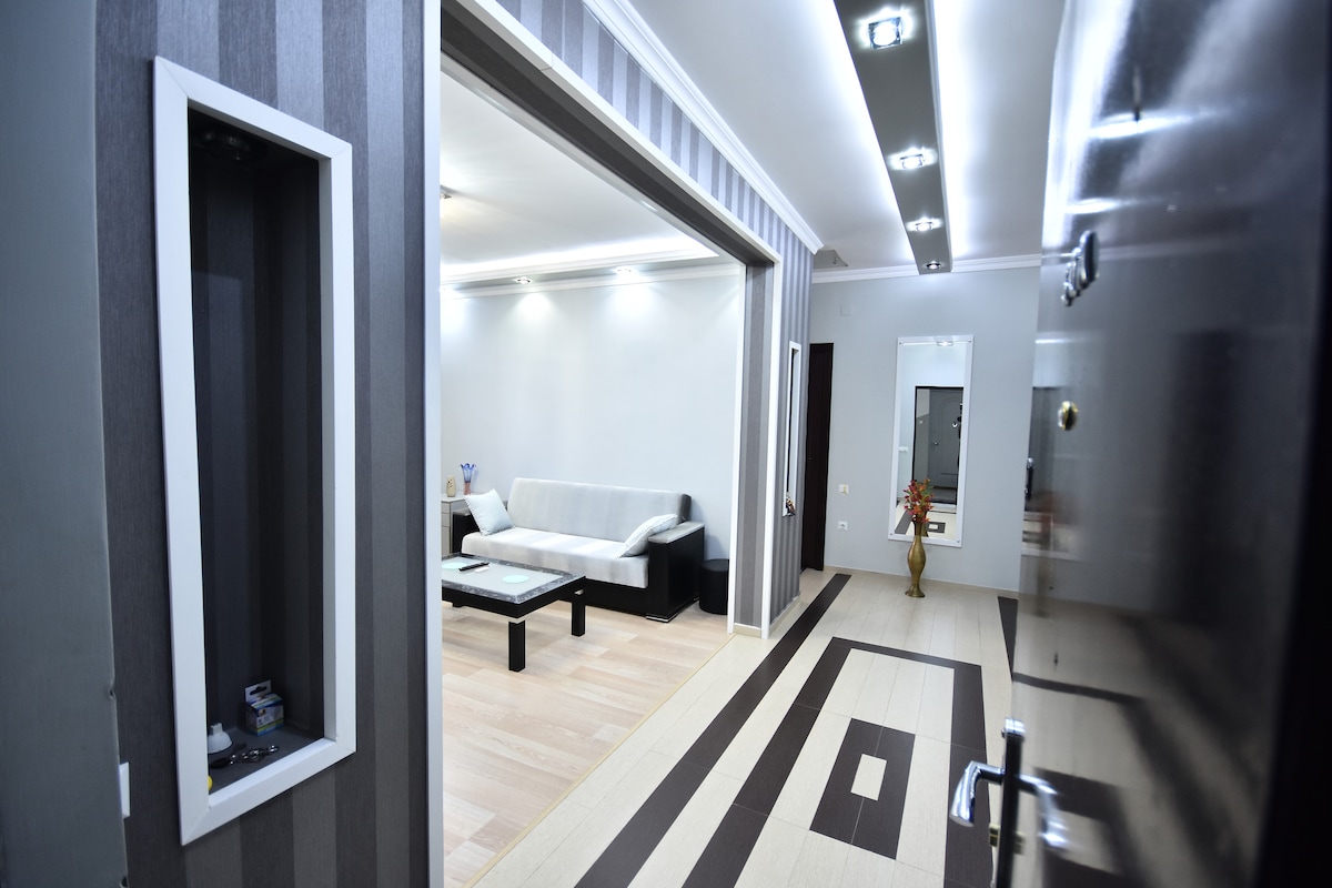 A hallway leads into a modern living area, featuring a light gray sofa and a black coffee table. Decorative elements are present in niche spaces. Soft lighting enhances the ambiance, and the floor displays a geometric pattern in neutral tones.