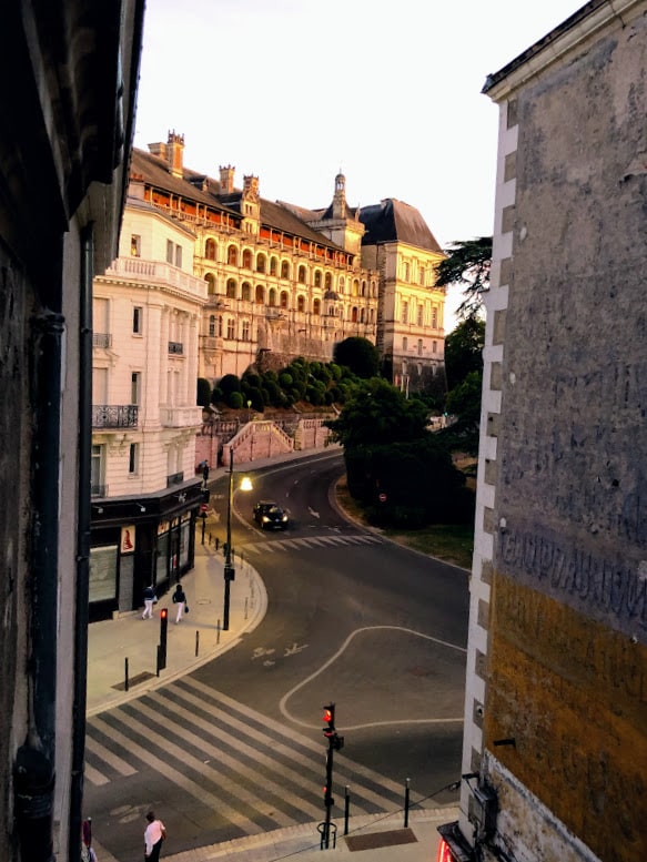 A view from a window displays the historical architecture of a grand building across the street, set against a twilight sky. The scene captures a serene road with a striped crosswalk and a traffic light, creating a dynamic yet peaceful urban environment.