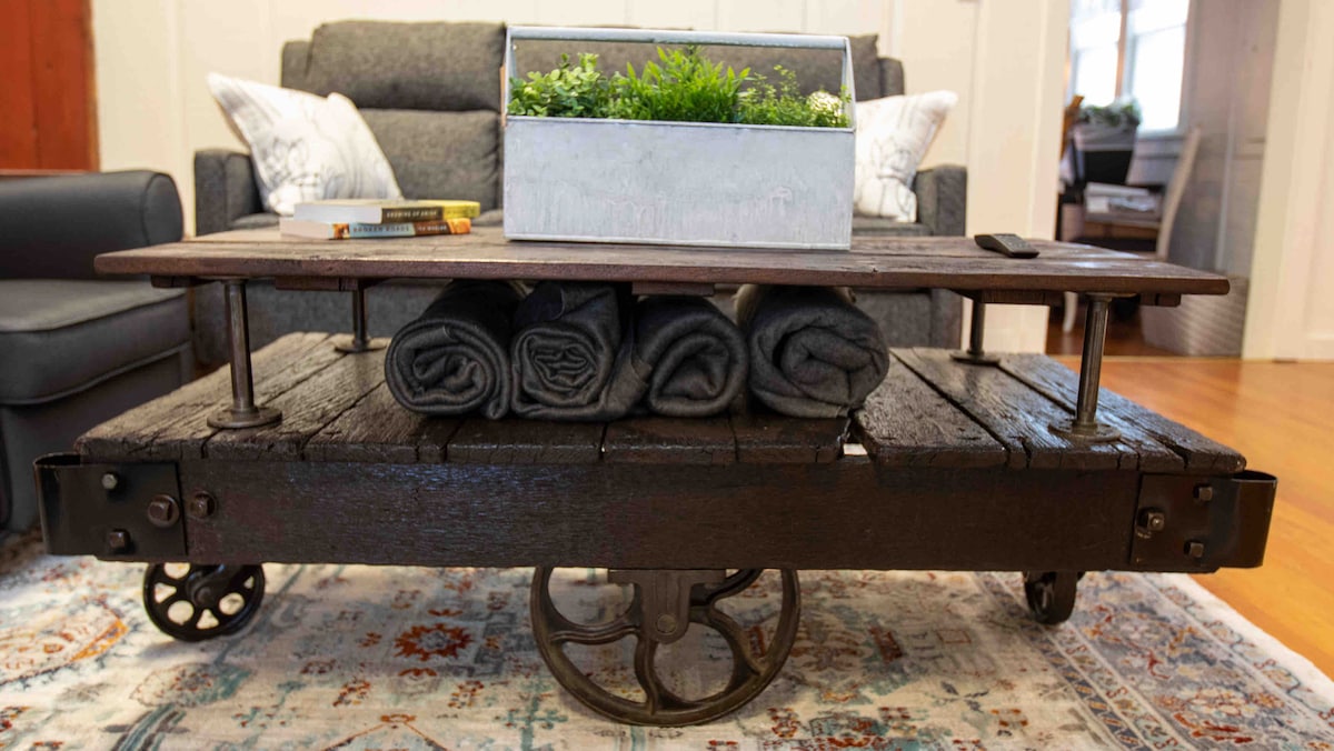 A rustic coffee table crafted from reclaimed wood is supported by sturdy metal wheels. Neatly rolled towels are stored beneath the table, accompanied by a small planter filled with greenery on its top surface. A comfortable seating area is visible in the background.