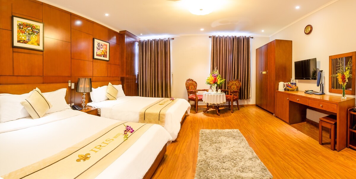 A spacious hotel room features two beds with neutral linens and decorative throws. A small seating area includes two chairs and a round table, illuminated by natural light from large windows with dark curtains. Wooden accents and artwork enhance the warm decor.