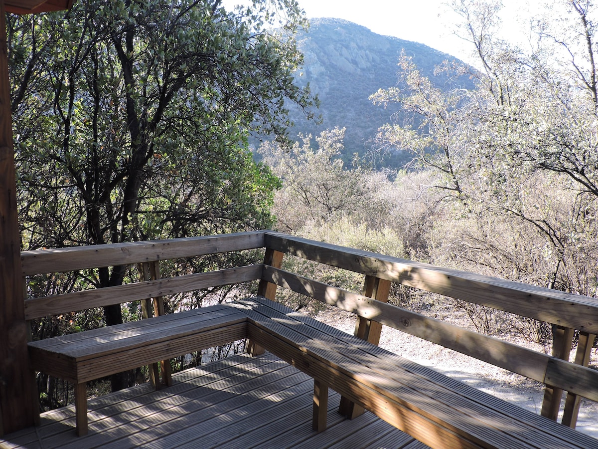 A wooden terrace with sturdy benches offers stunning views of the surrounding natural landscape. The area is framed by trees and shrubs, showcasing the tranquility of the environment with distant mountains visible in the background.