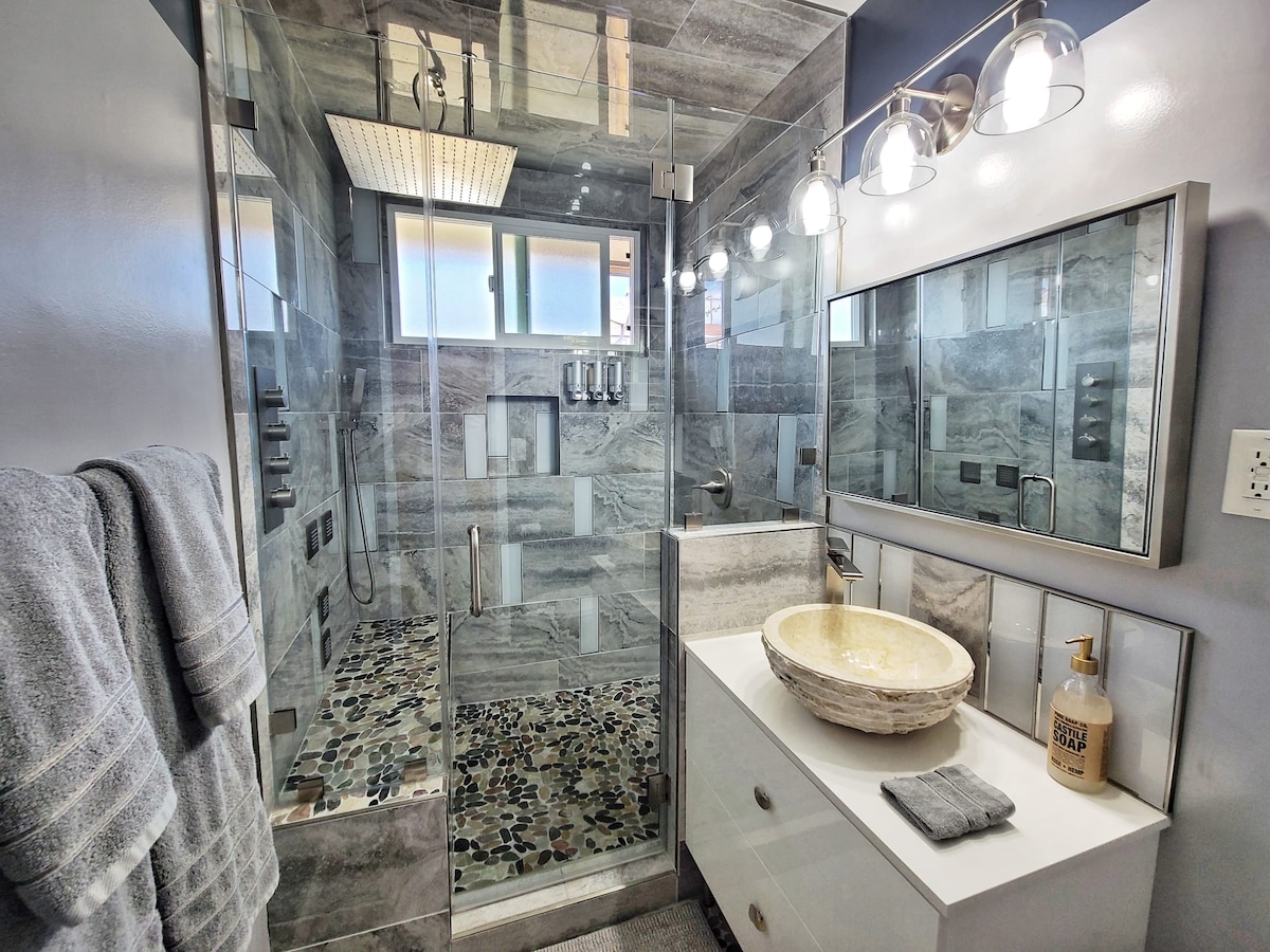 A modern bathroom features a glass-enclosed shower with gray stone tiles and a pebble floor. A stylish vessel sink sits atop a white vanity, accompanied by a soap dispenser. Soft towels are neatly hung near the shower, and ample light is provided by overhead fixtures.