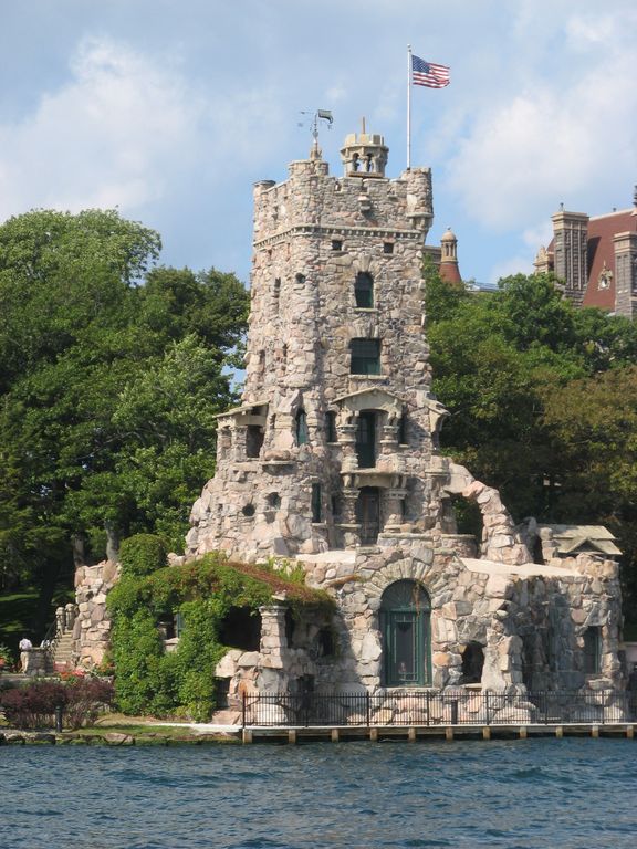 A historic stone tower stands along the water's edge, adorned with greenery. The structure features a flag waving above and intricate stonework. Surrounding trees complement the scenic view, which is framed by the blue water in the foreground.