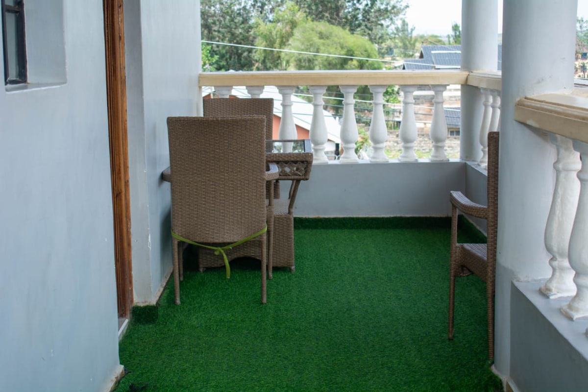 A small balcony features synthetic grass flooring, creating a cozy space. Two wicker chairs are positioned around a matching table, offering a spot for relaxation. The surrounding railing provides a view of the outdoors, enhancing the inviting atmosphere.