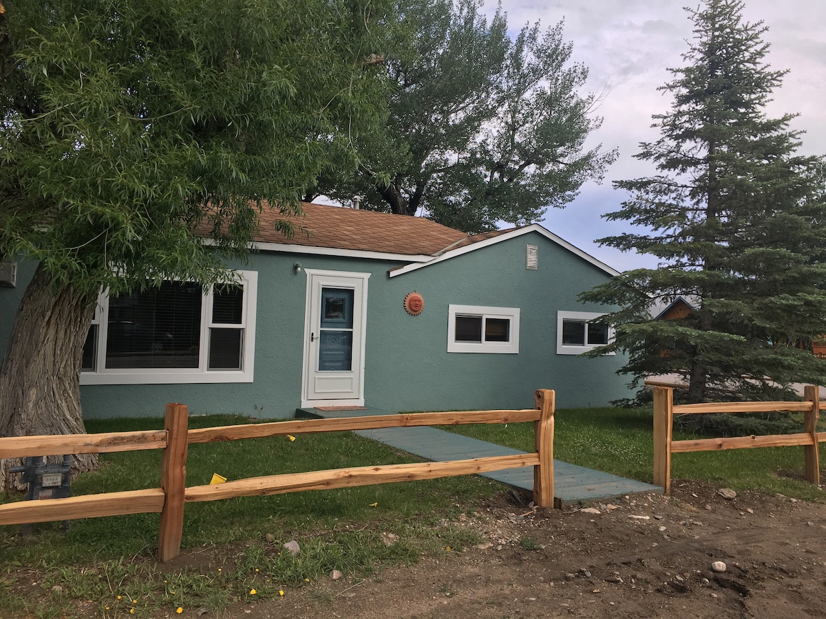 The cottage exterior features a light blue facade complemented by white trim. A pathway leads to the front door, with a wooden fence outlining the grassy yard. Mature trees provide shade, enhancing the inviting entryway.