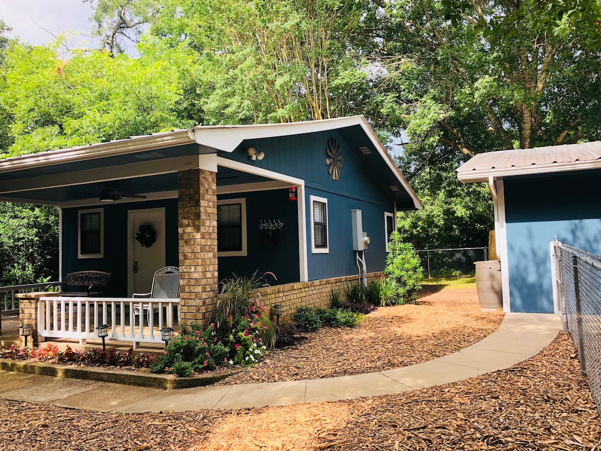 A charming cottage is nestled among trees, featuring a welcoming porch with seating. A flower bed with vibrant plants enhances the outdoor space. A paved path leads to a secondary structure, with a well-maintained exterior and a circular driveway providing easy access.