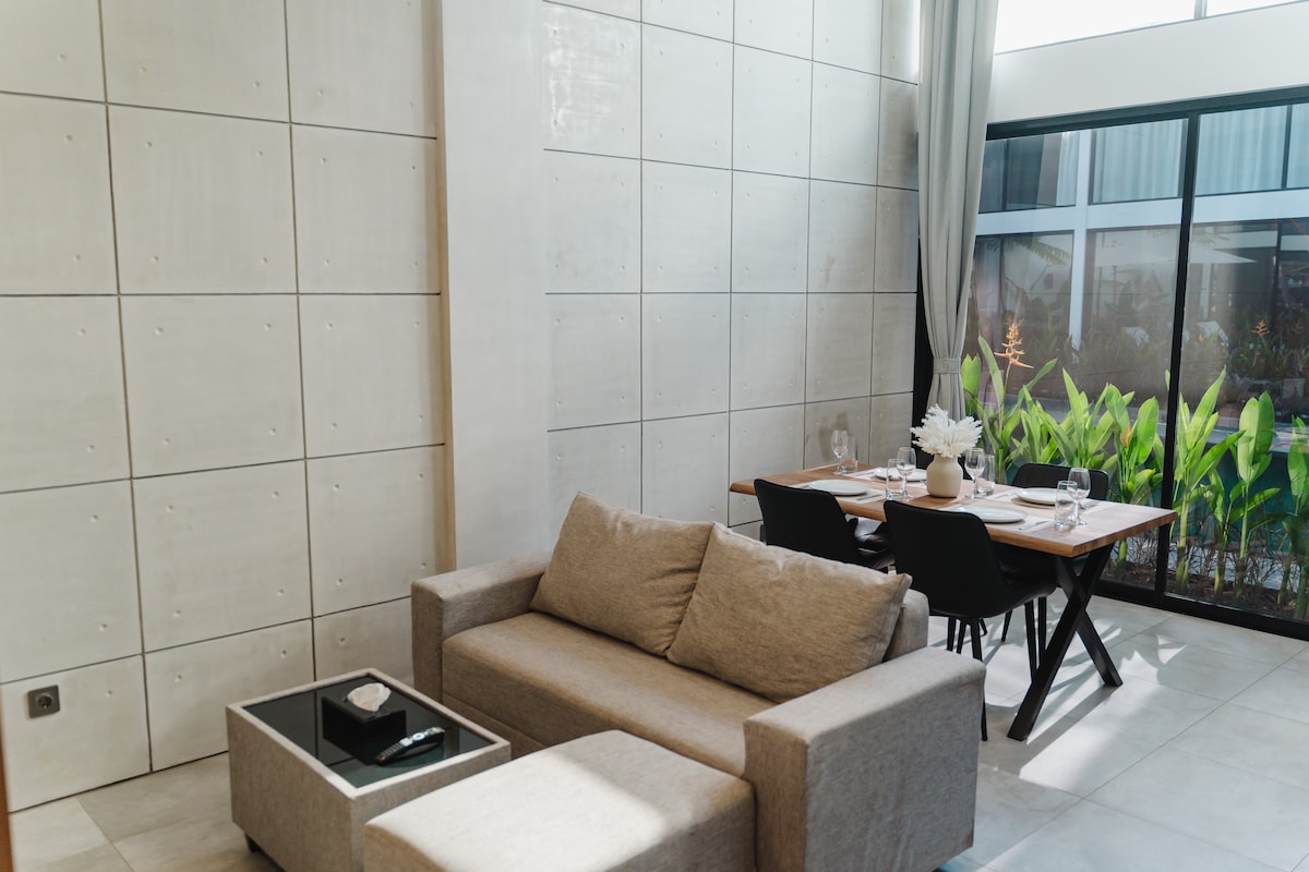 A modern living area is adorned with a soft gray sofa and a sleek coffee table, creating a comfortable space. A well-set dining table is positioned nearby, complemented by black chairs. Large windows allow natural light to fill the room, while greenery is visible outside.